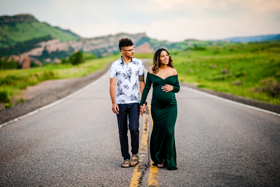 A couple walking on an empty road as she holds her belly in a fitting freen maternity dress