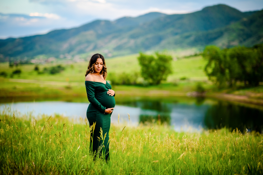 A woman in a fitting dark green maternity dress holding her belly at sunset in the foothills of Colorado