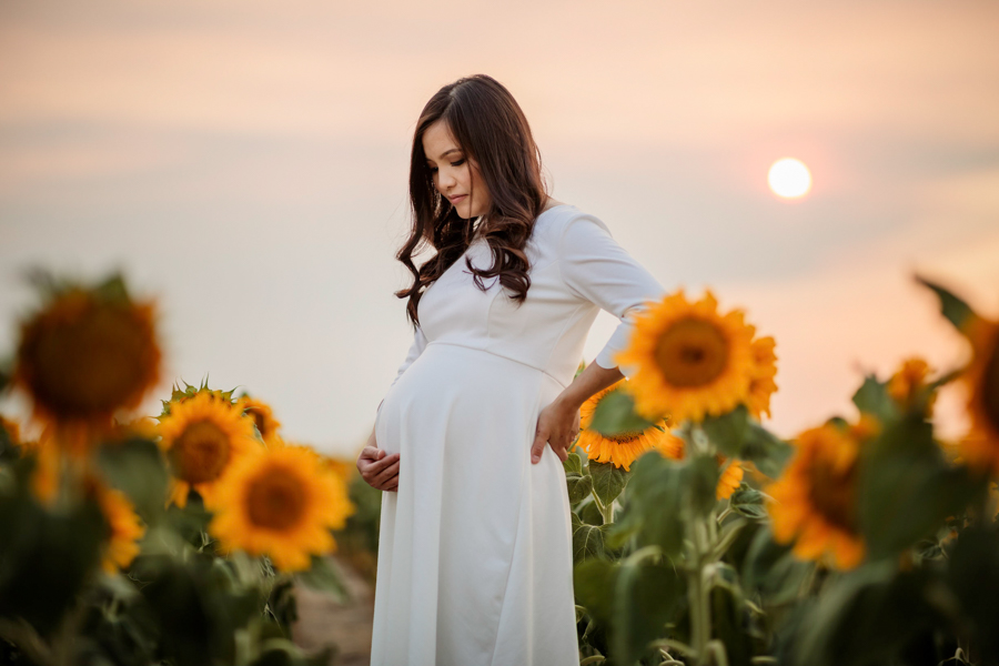 A woman in a white maternity dress holding her belly in sunflower fields at sunset