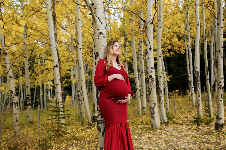 A woman in a fitting red maternity dress leaning on an apen tree in the fall