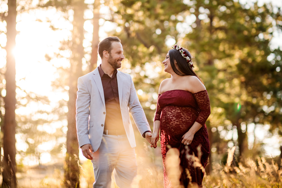 A couple holding hands in the woods while she wears a burgundy lace maternity dress