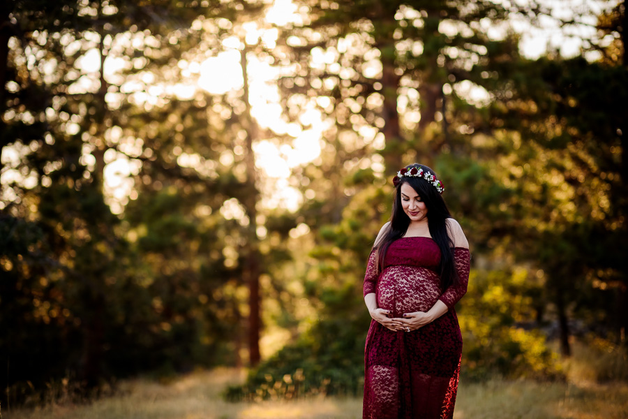 A woman in a burgundy lace maternity dress hugging her belly in the woods