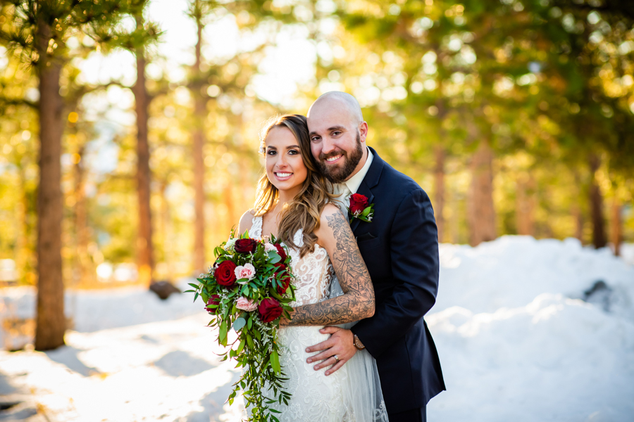 a newlywed couple smiling at the camera with trees and snow in the background