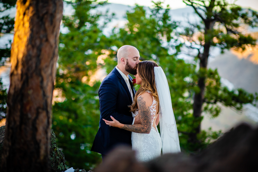 a newlywed couple standing on a mountain while the groom kisses her forehead