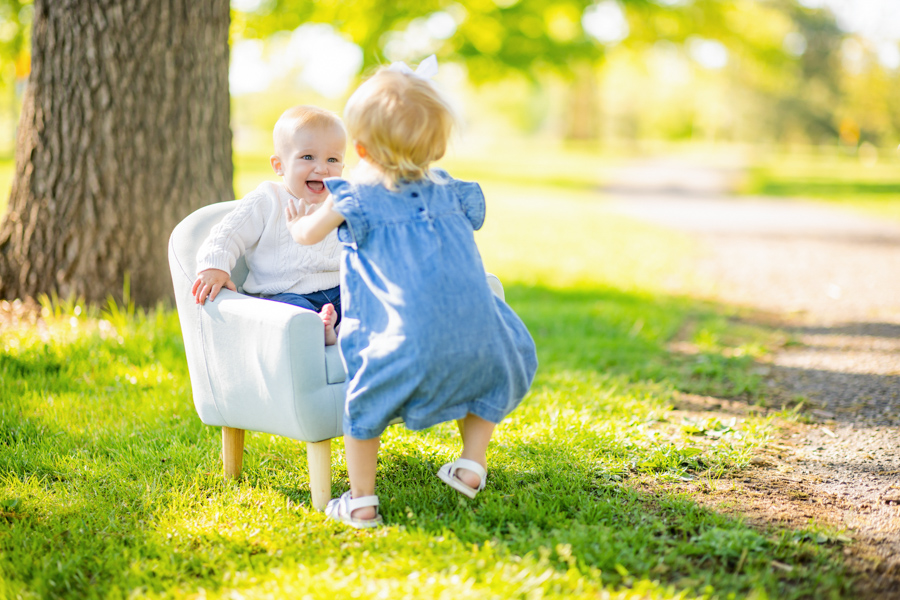 a one year old boy sitting on a chair laughing while his two year old sister tickles him